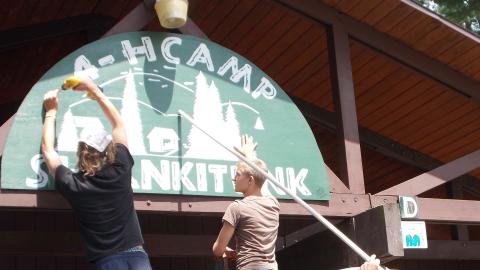 camp workers hanging up Camp Shankitunk sign, two people on ladders and one standing on the ground holding the sign with a pole