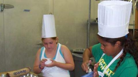 Two youth in aprons and chef hats work together to spoon batter into muffin tins.