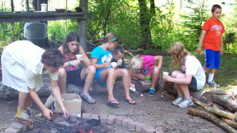 Group of campers gathered with cups of water around a fire pit while a counselor sets up a cooking grate over the fire.