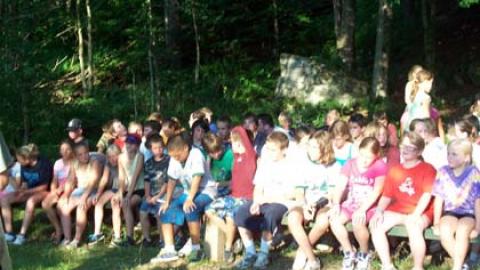 campers seated on benches in a wooded area watching a performance by a platoon group in front.