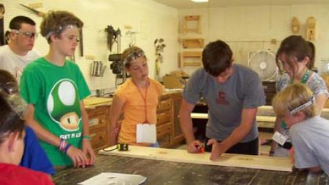 campers stand gathered around a table while counselor demonstrates measuring for a piece of the woodworking project.