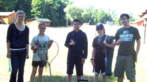 Two counselors stand with three youth and show off their fishing gear: nets, poles, and tacklebox.