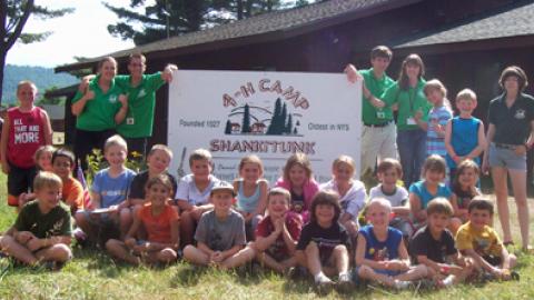 day campers sitting in front of camp shankitunk sign smiling with counselors in the back.