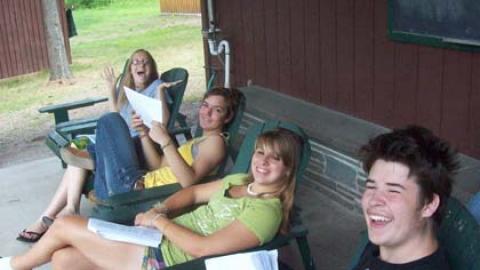 Teens in Adirondack chairs on front porch holding songbooks.