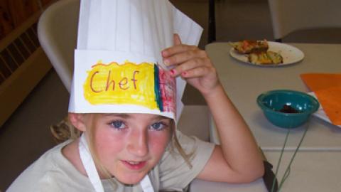 youth in apron and paper chef hat sitting over a plate of baked potatoes with extra veggies to create a mouse.