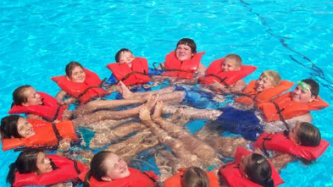 group of youth in the swimming pool wearing life jackets and linking arms to form an orange rescue circle.