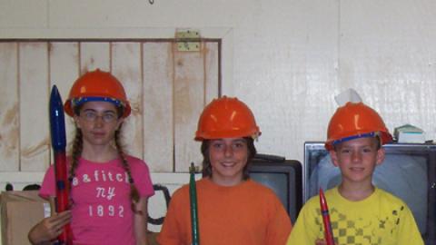 Three youth in hard hats holding model rocket kits.
