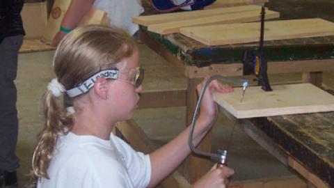 Youth kneels down to cut board with small hack saw in wood shop.