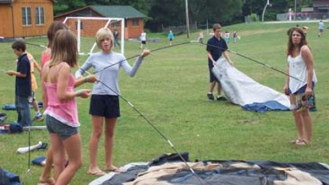 campers assembling tent poles to practice pitching a tent while counselor looks on making sour face at camera