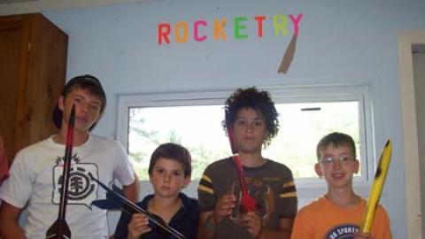 Four youth stand in front of Rocketry sign on wall holding model rockets they have built.