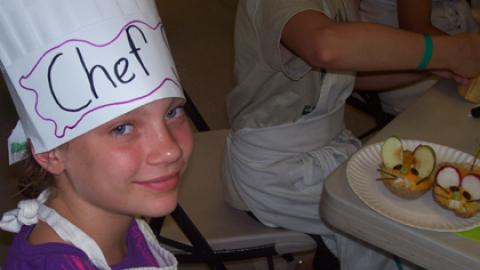 Youth in paper chef hat sitting near culinary creation.