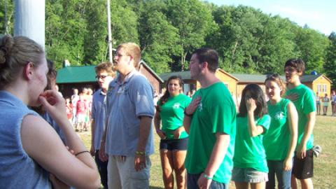 camp staff in matching green shirts saying the pledge of allegiance before lowering the flag while campers look on.
