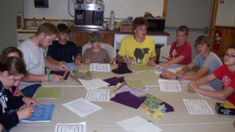 Campers seated around a table using paper pattern to create 9 mans morris boards on cloth using sharpie markers.