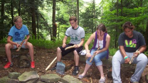 Youth sit on logs around a fire pit whittling.