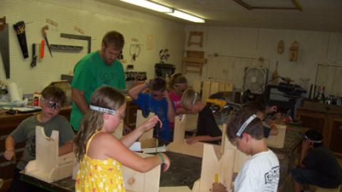 group of youth around a table with counselor in various stages of bench assembly using screw drivers and wearing goggles.