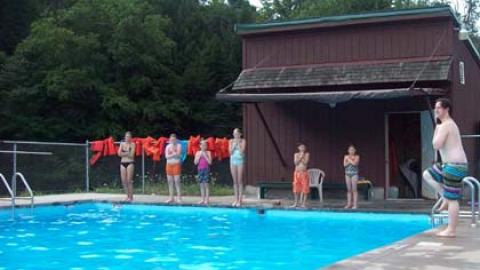 campers stand poolside while counselor demonstrates a kicking movement from the pool deck.
