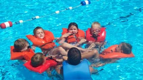 group of youth in the pool wearing life jackets and linking arms to form an orange rescue circle. 