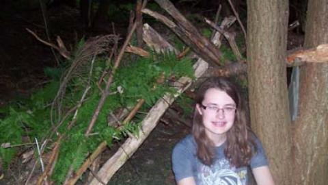 youth sitting in front of a shelter she built using sticks and ferns.