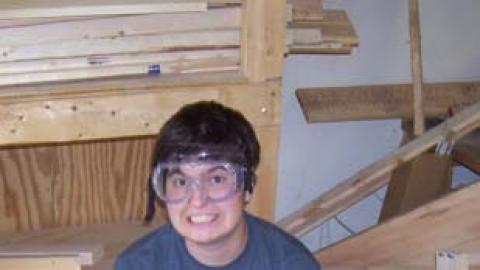 CIT II candidate sitting on bench in wood shop wearing safety goggles and writing notes on clipboard.