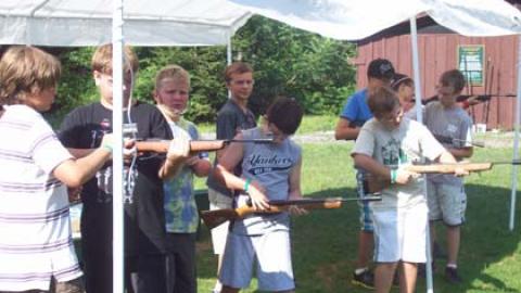 Five youth stand with air rifles at the line to fire while others assist their shooting partner or wait quietly behind the line for their turn.