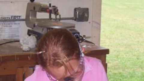 Young lady wearing safety goggles and sanding one leg of her woodworking project, a bench