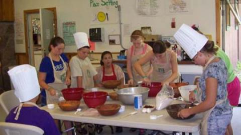 Campers gathered around a table with bowls and spatulas wearing paper chef hats and aprons.