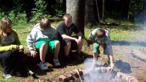 Youth set pie iron cookers in the fire pit as others are seated on logs around the pit.