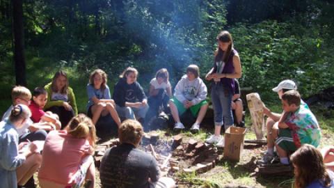 group of campers seated on logs around the fire pit with a counselor standing at the firepit.