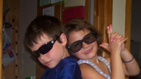 two young children stand back to back wearing sunglasses and holding their pointer fingers together.