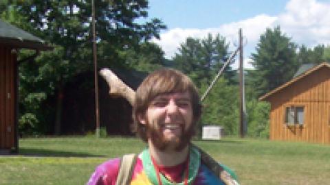 Smiling staff dressed in tie dye holding stick, radio, whistle, and lawn chair.