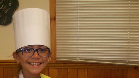Youth wearing a chef's hat stands in front of the food that is being cooked. 