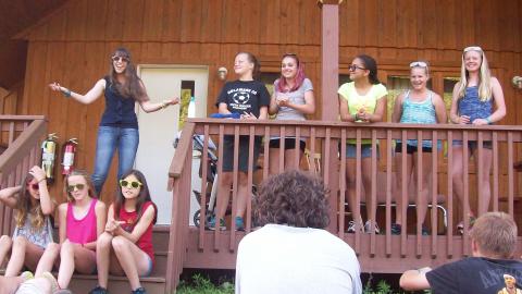 Campers and staff stand on directors porch during skit laughing and smiling down at audience on lawn.