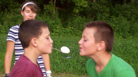 campers pass an egg between each other during a relay race where eggs are held on spoons in their mouths. A counselor watches in the background 