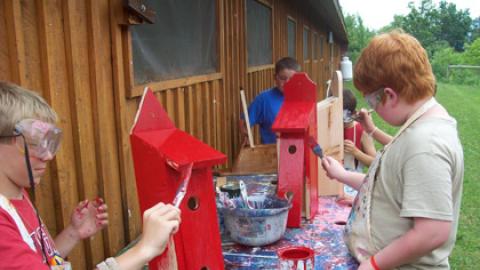 campers wearing aproms and goggles apples a coat of paint to their double-deck bird houses out behind the woodshop.