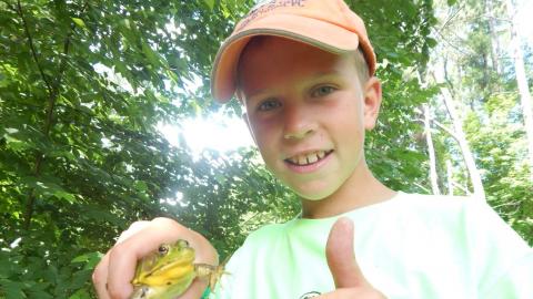 Camper holding a large frog and giving the camera a thumbs up in wooded area.
