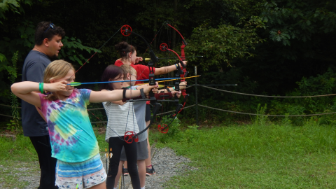 Archery youth stand on the firing line with their bows drawn prepared to fire as a counselor looks on over their shoulder.