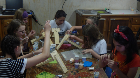 Campers seated around a table stringing beads onto wooden looms.