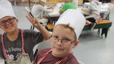 Two campers pose with their chef's hats. Youth in the back holds up two fingers for the camera. 
