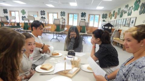 Youth sitting at a table laughing with paper directions as counselor attempts to pour jelly onto bread from large jar.
