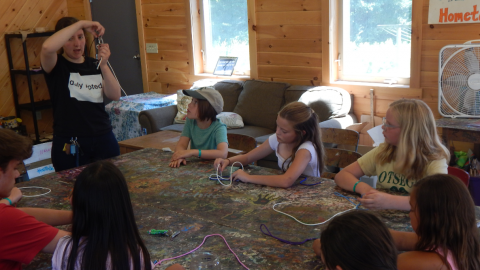 Craft Director demonstrates paracord weaving to a group of youth seated around a table with pieces of paracord.