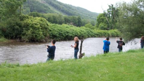 Youth stand along the bank of the Delaware River in various stages of casting and waiting for a bite during fishing class.