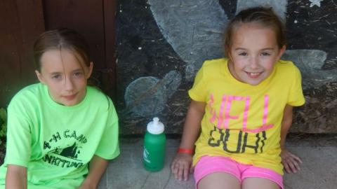 Two smiling campers sit on the cement with a smiley face necklace and water bottle on the ground in-between them. 