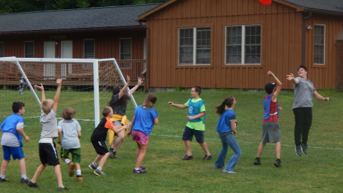 Counselor jumping into the air and throwing a frisbee over the heads of opposing team youth to those with arms raised to receive it.