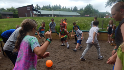 Youth scattered about in the gaga pit hitting a rubber ball toward each other while a counselor watches from outside the pit.
