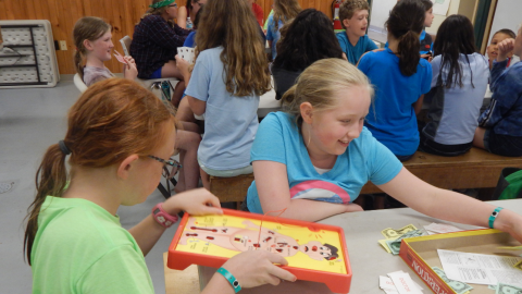 Youth at a table play operation. Others campers are seated at a table in the background playing other games.