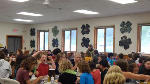 Campers sit at tables in the dining hall and eat a meal. 