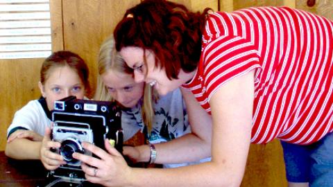 two youth examine an old fashioned camera with guidance from their smiling class counselor.