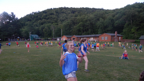 Youth with painted face making hand gesture toward camera as camp-wide game is played in the background.