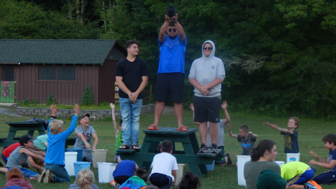 Counselor standing atop a picnic table holding a toy gorilla with campers below bowing to it.