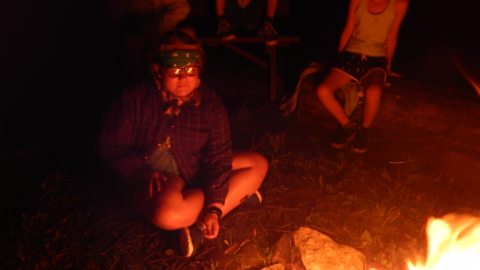 Youth seated in front of the campfire looking into the flames with others seated on a log behind them.